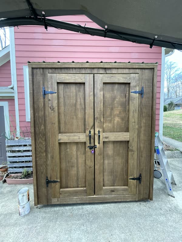Custom stained-pine storage cabinet with wrought-iron strap hinges against a red clapboard house